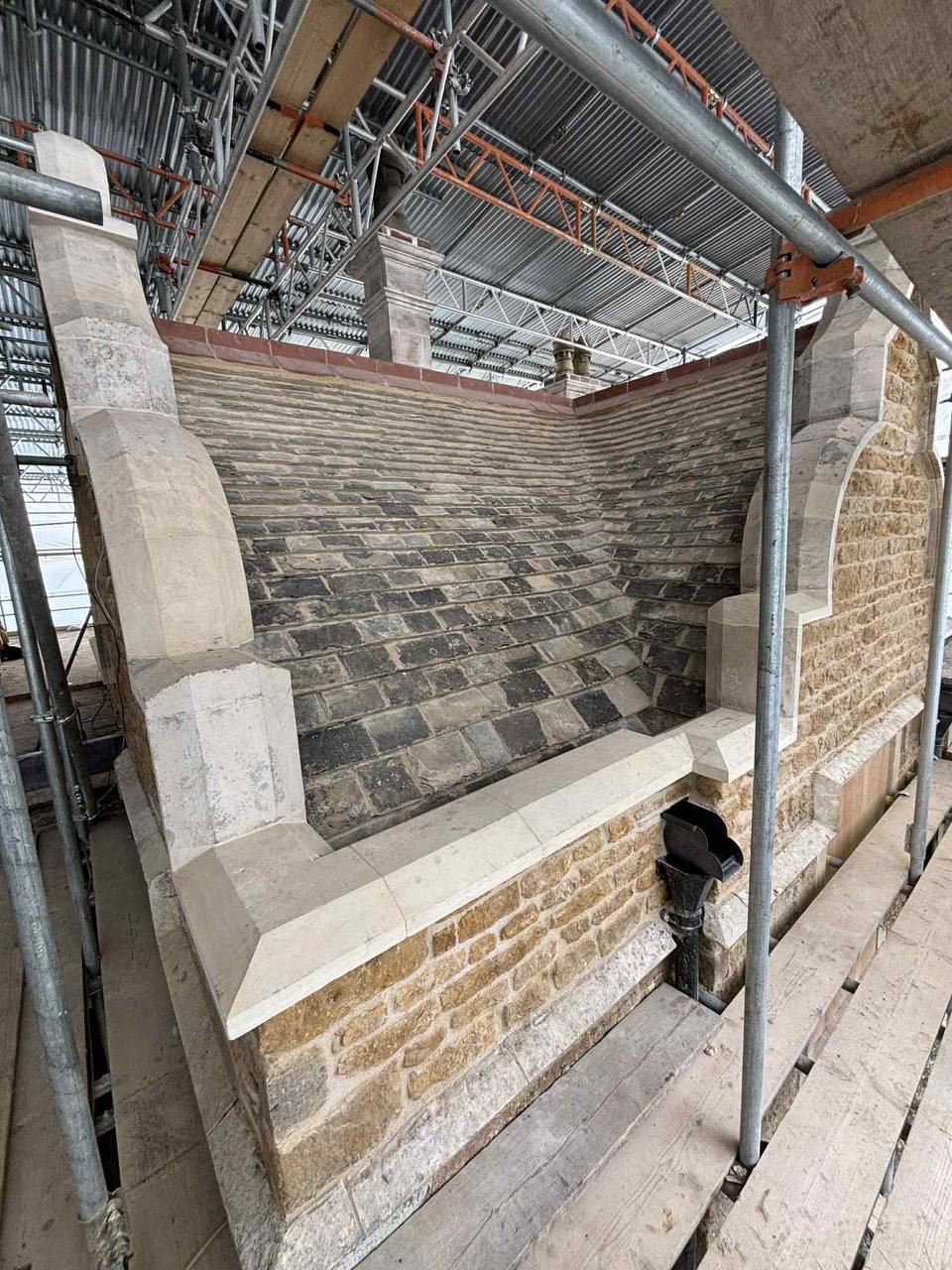 Restored stone parapet gutter and coursed stone-slate valley behind a shaped ironstone gable, with new dressed limestone copings during conservation works